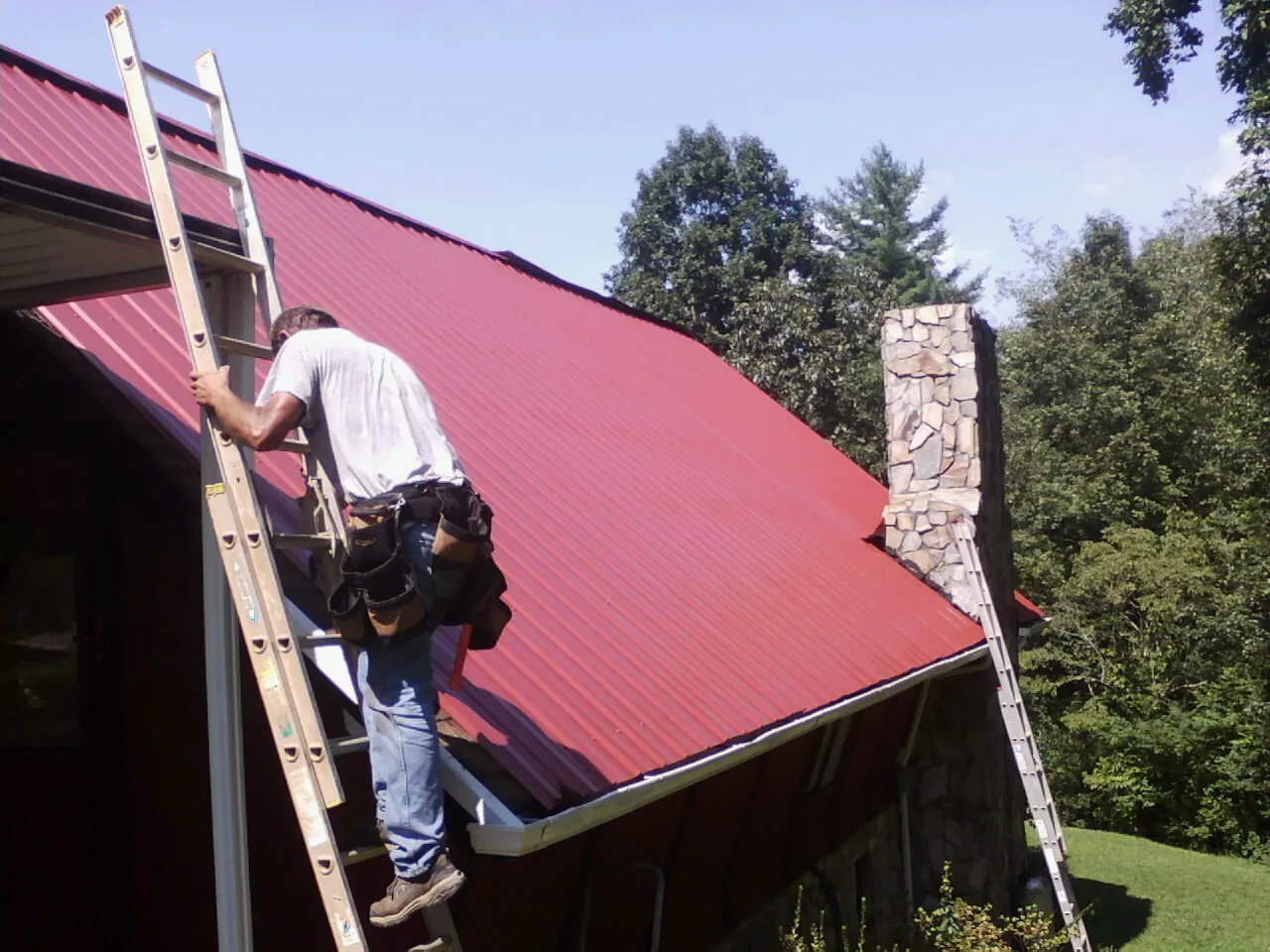 Roofer on a ladder beside a red metal roof and stone chimney — Soffit & Fascia Repair by Roofing Brothers in Sammamish